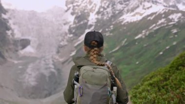 Sırt çantalı bir kadın Svaneti dağlarında yürüyüş yapıyor. Jangi-Tau buzulu manzarası, yeşil bitkiler. Dağlarda tek başına yürüyüş yapan biri. Macera, doğa keşfi, doğa turizmi. Çoklu gün yürüyüşü. Yavaşla