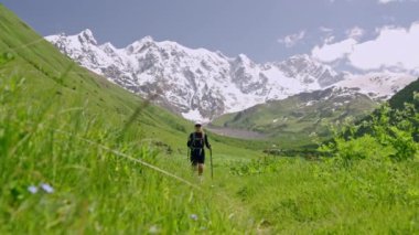 Jangi-Tau vadisi Svaneti 'de sırt çantalı kadın turist yürüyüşleri. Kadın yeşil tepelerin ve açık mavi gökyüzünün altındaki karlı dağların arasında yürüyüş direkleriyle doğa izlerini araştırıyor. Yavaş çekim.
