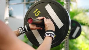 Formda bir birey, antrenman ekipmanlarının güç eğitimi için sabitlenmesini sağlar. Güneşli açık spor salonunda güvenli bir şekilde taşımak için halterde elleri sıkı bir çene kilidi var. Ağırlık hazırlıyorum, sporda güvenliğe odaklanıyorum. Yavaşla