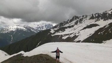 Solo sırt çantalı adam yüksek karlı dağlarda duruyor. Hava görüntüsü. Karla çevrili bir yürüyüşçü, engebeli tepeler, bulutlu gökyüzü. Açık havada macera gezintisi, doğa keşfi muhteşem manzara dronu.