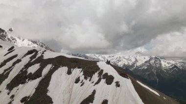 Karla kaplı tepeleri ve bulutlu gökyüzü olan Svaneti dağlarının hava manzarası. İki yürüyüşçü ıssız bir tepede yürüyor. Çarpıcı manzara seyahat, macera, doğa ve turizm için mükemmel..