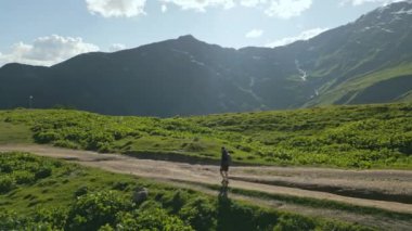 Dağlık çayırlarda gezen sırt çantalı adamın hava manzarası. Yeşil tepeler, dağ tepeleri ve açık mavi gökyüzüyle çevrili toprak yolda yürüyen biri. Açık hava macerası, seyahat doğa keşfi. İHA atışı