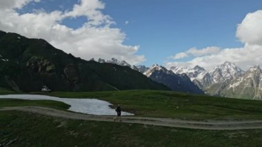 Yürüyüşçü yüksek dağlarda dağlık çayırlarda yürür. Yeşil tepelerde kar yamaçları. Mavi gökyüzünün altında bulutlu görkemli zirveler. Açık hava macerası, doğa keşfi. Manzaralı yaz yürüyüşü rotası.