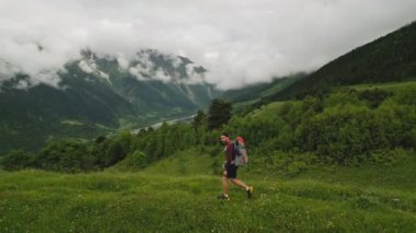 Yeşil bitkilerle, karla kaplı dağlarla, bulutlu bir günde nehir akıntısıyla dağlarda sırt çantasıyla gezen bir adam. Açık hava macerası, dağlık arazide yürüyüş. Yaz seyahati doğa keşfi.