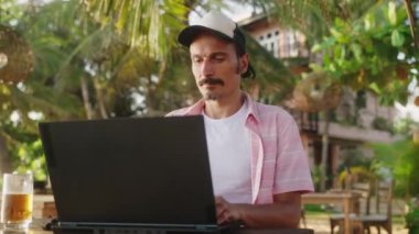 Freelancer ekrana odaklanıyor, palmiye ağaçları ve deniz meltemi ile uzak bir işi seviyor. Günlük kıyafet giyen adam tropik plaj kafesinde dizüstü bilgisayarda çalışıyor. Masada bira bardağı, güneş ışığında dijital göçebe yaşam tarzı. Yavaşla