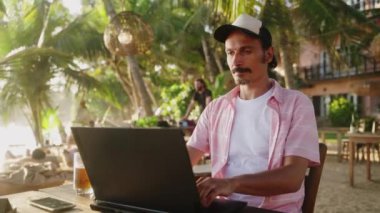 Freelancer ekrana odaklı, dijital göçebe konsepti, güneşli sahil çalışma alanı. Dizüstü bilgisayarı olan adam tropikal plaj kafesinde çalışıyor, palmiye ağaçları altında uzak iş hayatı yaşıyor. Yavaş çekim.