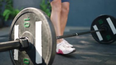 Dövmeli bacaklar gücü gösteriyor, kaldırmadan önce odaklan. Atletik kadın halterci açık hava spor salonunda ağırlık kaldırmaya hazırlanıyor, halteri kavrıyor. Spor elbiseli kadınlar, spor ayakkabılar, bilek sargıları....