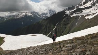 Sırt çantalı, yüksek dağlarda tek başına yürüyen adamın hava manzarası. Karla kaplı tepeleri ve yeşil vadileri olan dramatik bir manzara. Macera, keşif, doğa, doğa, doğa yürüyüşü. İHA atışı