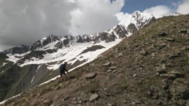 Adam sırt çantasıyla yüksek dağlara tırmanıyor. Solo dağcı kayalık yamaçta yürüyüş yapıyor. Karlı tepelerin havadan görünüşü. Açık hava macera sporu. Uzak vahşi doğa. İHA atışı