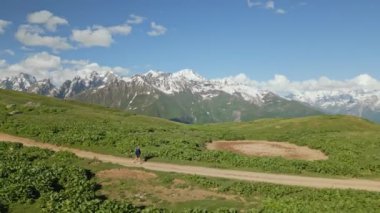 Yalnız gezgin, Svaneti dağlarındaki dağlık çayırlarda toprak patika boyunca yürüyor. Mavi gökyüzünün altında kar kaplı tepeleri olan yeşil manzaranın havadan görünüşü. Doğa macerası yürüyüş deneyimi. İHA atışı