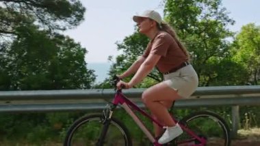 Aktif dişi pembe bisikletle doğayı keşfediyor, sağlıklı yaşam tarzı macerası. Genç kadın kozalaklı ormanda bisiklet sürüyor, güneşli bir günde deniz manzarasından hoşlanıyor..