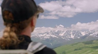 Yürüyüşçü kadın, Büyük Kafkasya 'daki Svaneti dağlarının panoramik manzarasını seviyor. Sırt çantası, şapka ve güneş gözlüğü takıyordu. Yeşil tepeler ve karlı tepeler. Yaz güneşli bir gün. Yavaş çekim.
