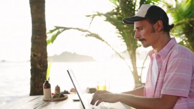 Freelancer günbatımını, yazarken deniz manzarasını, doğada iş-yaşam dengesini sever. Adam sahildeki kafede dizüstü bilgisayarla çalışıyor. İçkili, tropikal ortamda serbest çalışıyor. Yavaş çekim.