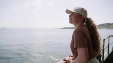 Gülümseyen kadın gezgin güneşli bir günde rahatlar, uzak manzarayı izler. Kadınlar tekne korkuluklarından deniz manzarasını sever. Macera turizmi, tek başına seyahat, okyanus manzarası..