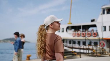 Kadın yolcu rıhtımda, güneşli deniz kıyısında, arka planda su araçlarıyla kalkış için bekliyor. Feribota binen kadın, uzun saç, günlük kıyafet, güneşli rıhtım manzarası..