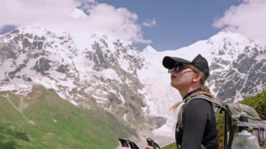 Sırt çantalı kadın yürüyüşçü, direkler dağ manzarasını sever. Svaneti dağlarında karlı tepelere karşı duruyoruz. Açık hava macerası, doğa, yürüyüş. Güneşli bir gün, berrak bir gökyüzü, tırmanma yolculuğu.