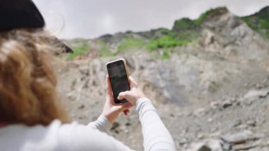Kadın yürüyüşçü dağlık alanda fotoğraf çekmek için telefon kullanıyor. Gezici kayaların ve yeşil tepelerin manzaralı görüntülerini yakalar. Dağlık alanlarda doğa fotoğrafçılığı. Yavaş çekim.