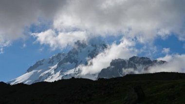 Kafkasya sırtındaki Kazbegi Dağı 'nın zamanı. Bulutlar karlı tepenin etrafında hareket ediyor. Yüksek dağlarda değişen hava koşullarıyla görkemli bir manzara. Açık mavi gökyüzü ve kayalık arazi görünür..