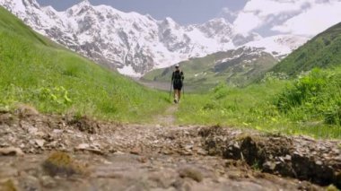 Svaneti 'deki Jangi-Tau vadisinde sırt çantalı bir turist. Gezgin doğa yürüyüşleri, direklerle ve dağ manzaralarıyla gezer. Arka planda karla kaplı zirveler. Yavaş çekim.