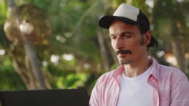 Serbest çalışan tipler, palmiye ağaçları ve deniz meltemi arasında uzak bir işi seviyor. Şapkalı adam tropik plaj kafesinde dizüstü bilgisayarla çalışıyor. Dijital göçebe yaşam tarzı, güneşli okyanus kıyısında iş hayatı dengesi. Yavaşla