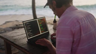 Erkek yazma, dizüstü bilgisayarda kod düzenleme, tropikal deniz manzarası. Günbatımında Oceanfront Beach Cafe 'de web geliştirici programları. Uzaktan çalışma, rahatlatıcı sahil atmosferiyle birleşir. Yavaş çekim.