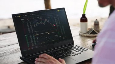 Erkek monitörler kripto para biriminde dalgalanmalar, okyanus kıyısında online yatırımlar. Kripto tüccarı, Sunset Beach Cafe 'de dizüstü bilgisayardaki pazar haritalarını analiz ediyor. Serbest çalışanların arka planı, deniz melteminin tadını çıkarıyor..