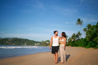 Erkek ve kadın deniz kenarında çıplak ayakla yürüyor, tatilin tadını çıkarıyorlar. Güneşli bir günde tropik plajda mutlu çift gezintileri. Deniz kenarında romantik bir yürüyüş, aşk, boş zaman. Gezginler kıyı manzarasını keşfeder.