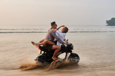 Couple rides motorbike on sandy beach. Man drives motorcycle while woman spreads arms. Lovers enjoy romantic beach, vacation at sunset. Adventure and freedom vibes. clipart