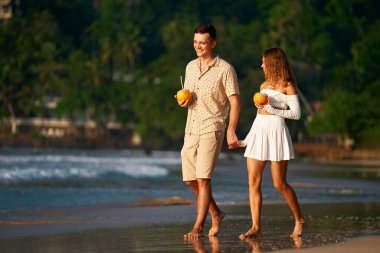 Pair with tropical drinks walk by sea, honeymoon vacation mood on exotic island getaway. Romantic couple stroll barefoot on sandy beach, man and woman in summer clothes hold hands enjoying sunset. clipart