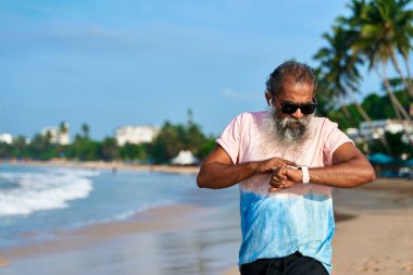 Sahilde kıdemli Güney Asyalı koşucu. Akıllı, kablosuz kulaklıkları var. Sakallı adam sabah koşusu sırasında spor takip cihazını kontrol ediyor. Sağlık için modern teknoloji. Aktif yaşam tarzı olgun fitness meraklısı.
