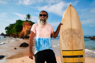 Son sınıf sörfçü elinde sörf tahtasıyla kumlu sahilde duruyor. Gülümseyen yaşlı erkek aktif emekliliğin tadını çıkarıyor. Okyanus dalgaları kıyıya çarpar. Mutlu altın yıllarda sörf yapmak. Yaşam tarzı, özgürlük macerası.