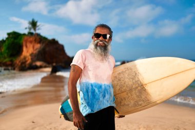Sunny Beach 'te sörf tahtası olan son sınıf öğrencisi. Yaşlı sörfçü gülümser, güneş gözlüğü takar. Aktif yaşam tarzı, okyanus dalgaları arka planı. Deniz kenarındaki kıyı macera fitnesinin tadını çıkar. Mutlu emeklilik.