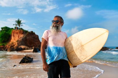 Sunny Beach 'te son sınıf siyah adam sörf tahtası tutuyor. Aktif yaşlı sörfçü okyanus dalgalarına gülümsüyor, sörf yaşam tarzını seviyor. Yaşlanan erkek macerayı kucaklıyor, canlı bir ruh sergileyen sağlıklı bir deniz yaşamı..