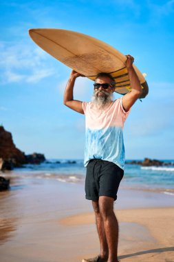 Aktif kıdemli adam güneşli sahilde sörf tahtası tutuyor. Şık giyinmiş etnik erkek, sörf yapıyor. Yaşlı sörfçüler denizde macera gülümsemelerinden hoşlanırlar. Sahilde sağlıklı bir yaşam tarzı. Seyahat siteleri için resim.