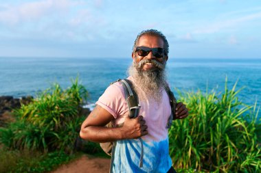 Sırt çantalı gülümseyen yaşlı adam okyanus manzarasının yanında duruyor. Güneş gözlüklü sakallı gezgin güneşli bir günde doğa macerasının tadını çıkarıyor. Üst düzey erkek keşif, sahil yolu. Açık havada seyahat yaşam tarzı.
