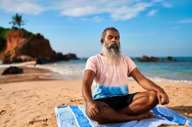 Yaşlı adam okyanus kıyısındaki kumlu sahilde meditasyon yapıyor. Çizgili mindere bağlanmış, gözleri kapalı, sakin, rahat oturuyor. Huzurlu kıyı yogası, mavi gökyüzünün altında farkındalık seansı. Doğa geri çekilirken zihinsel sağlık.