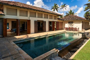 Döşeme çatılı ve yüzme havuzlu klasik tropikal villa. Palmiye ağaçları, bahçe mobilyaları. Güneşli bir gün, açık gökyüzü. Egzotik ortamda rahatlama ve sükunet. Tatil ya da dinlenme için mükemmel.