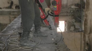 Tozla dolu hava, yenileme sürecinde beton kalıntıları. İnşaat işçileri hasarlı duvarları yıkmak için matkap kullanıyorlar, savaştan etkilenmiş apartmanın tavanlarını. Koruyucu giysili işçiler çalışıyor..