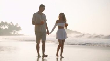 Romantik bir adam, deniz sörfünde el ele yürüyen bir kadın. Güzel bir çift gün batımında okyanus kıyısında rahatlayacak. Erkek arkadaş, kız arkadaş hindistan cevizi kokteyli içer, kumsalda buluşmanın tadını çıkarır..