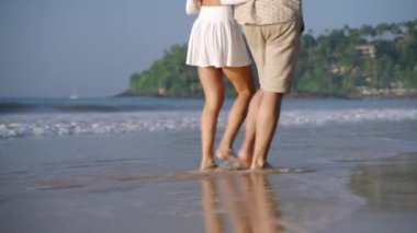 Dişi, erkek ayakları deniz dalgalarında yalınayak geziyor. Kumlu sahilde yürüyen insan bacakları. Genç çift romantik bir randevuda el ele tutuşurken konuşur. Erkek arkadaş, kız arkadaş tatilde rahatlar. Düşük açı.