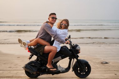Plajda birkaç motosiklet. Erkek motosiklet sürüyor, kadın ona sarılıyor, deniz manzarasının tadını çıkarıyor. Romantik macera, okyanus kıyısında seyahat eğlencesi. Günbatımı, tasasız aşk tatil özgürlüğü.