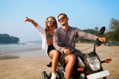 Kumsalda motosiklet süren bir çift. Adam gülümseyerek scooter kullanıyor. Kadınlar deniz ufkunu göstererek tropikal tatilin tadını çıkarıyorlar. İyi yolculuklar. Güneş parlıyor, okyanus manzarası.