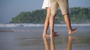 Dişi, erkek ayakları deniz dalgalarında yalınayak geziyor. Kumlu sahilde yürüyen insan bacakları. Genç çift romantik bir randevuda el ele tutuşurken konuşur. Erkek arkadaş, kız arkadaş tatilde rahatlar. Düşük açı.