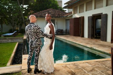 Tropikal villa 'da damadın yanında beyaz gelinlik içinde, cinsiyeti belirsiz, değişken bir insan. Seçkin bir çift, bilardo yakınlarındaki eşsiz törenleri kutluyor. Modayı takip eden gay düğün modası.