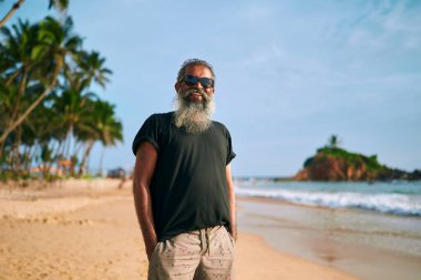 Yaşlı sakallı adam güneşli bir sahil gezisinin tadını çıkarıyor. Şık bir şekilde güneş gözlüğü takmış. Okyanus dalgaları arasında rahatça dolaşmak. Aktif kıdemli yaşam tarzı. Macera arayan emekliler için popüler seyahat yeri.
