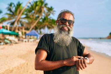 Kumsalda gri sakallı yaşlı adam akıllı saat kullanıyor. Güneş gözlüklü şık olgun erkek teknolojik aletlerden hoşlanır. Aktif yaşlı yaşam tarzı, giyilebilir açık havada yenilik. Arka planda okyanus palmiyeleri.