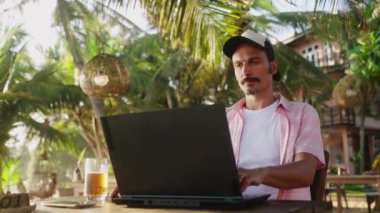 Dijital göçebe tipler, biralarını yudumluyor, palmiye ağaçları sallanıyor. Freelancer, tropik plaj kafesinde dizüstü bilgisayarda çalışıyor. Cennette sıradan bir iş hayatı. Günbatımı ışığı yaprakların arasından süzülür. Yavaş çekim.