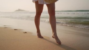 Kadın şapkasını tutuyor, deniz kenarında gezmekten hoşlanıyor, deniz kenarında özgürlükten zevk alıyor. Çıplak ayaklı kadın güneşli sahilde yürüyor, dalgalar ayaklarını yıkıyor. Tatil, tatil havası, yaz tatilleri..
