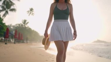 Bağımsız kadın sakin deniz manzarasını seviyor, şapkasını tutuyor. Solo kadın gezgin güneş doğarken tropikal sahilde yürür. Yolculuk tutkusu, okyanus kenarında yalnızlık anları. Gündoğumu yürüyüşü, huzur arayışı.