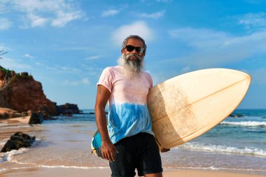 Kıdemli etnik adam okyanus sahillerinde sörf yapıyor, sörf tahtası tutuyor. Aktif yaşam tarzı, sağlıklı yaşlanma. Yaşlı erkek macera ve özgürlükten hoşlanır. Güneşli bir gün, deniz kenarında dinlenmek, zinde kalmak, pozitifliği kucaklamak..